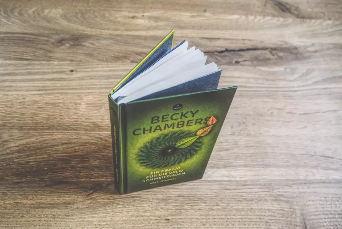 Ein Buch mit dem Titel „Becky Chambers – Ein Psalm für die Wildnis der Welten“ mit einem grünen Einband, auf dem ein Auge abgebildet ist, steht aufrecht auf einem Holztisch.