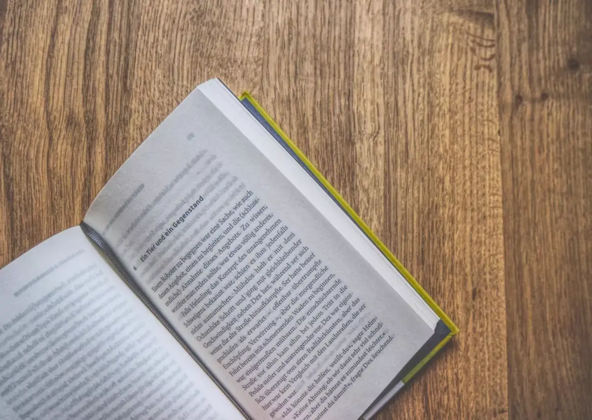 Ein offenes Buch mit sichtbarem Text, das auf einer Holzoberfläche liegt und dessen Seiten in der Nähe des Buchrückens leicht nach oben gebogen sind.