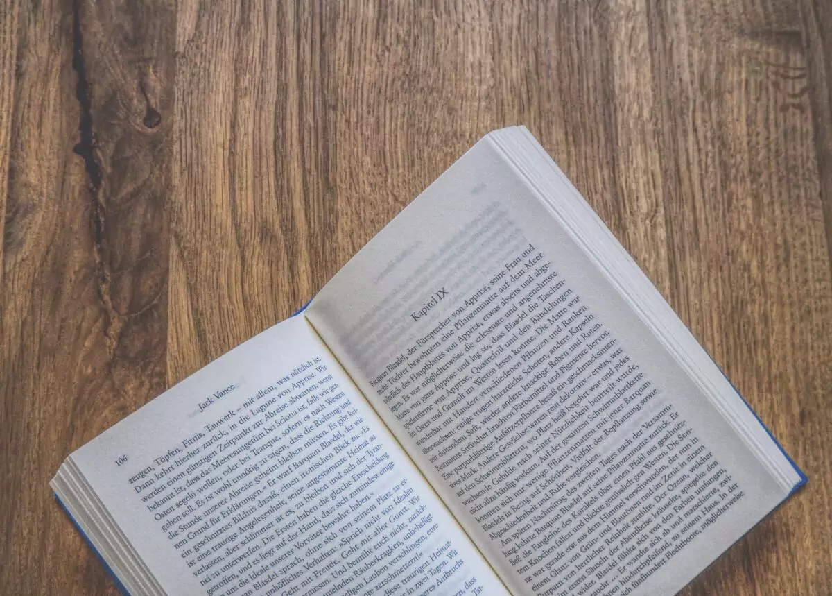 Auf einer Holzoberfläche liegt ein offenes Buch, das auf beiden sichtbaren Seiten Text anzeigt.
