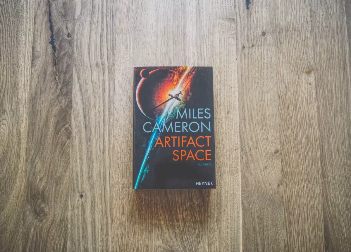 Ein Buch mit dem Titel „Artifact Space“ von Miles Cameron liegt auf einer Holzoberfläche.