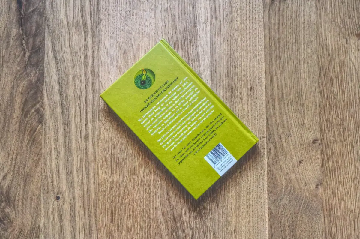 Ein grünes Taschenbuch, das mit der Vorderseite nach unten auf einer Holzoberfläche liegt. Auf der Rückseite sind Text und ein Strichcode zu sehen.