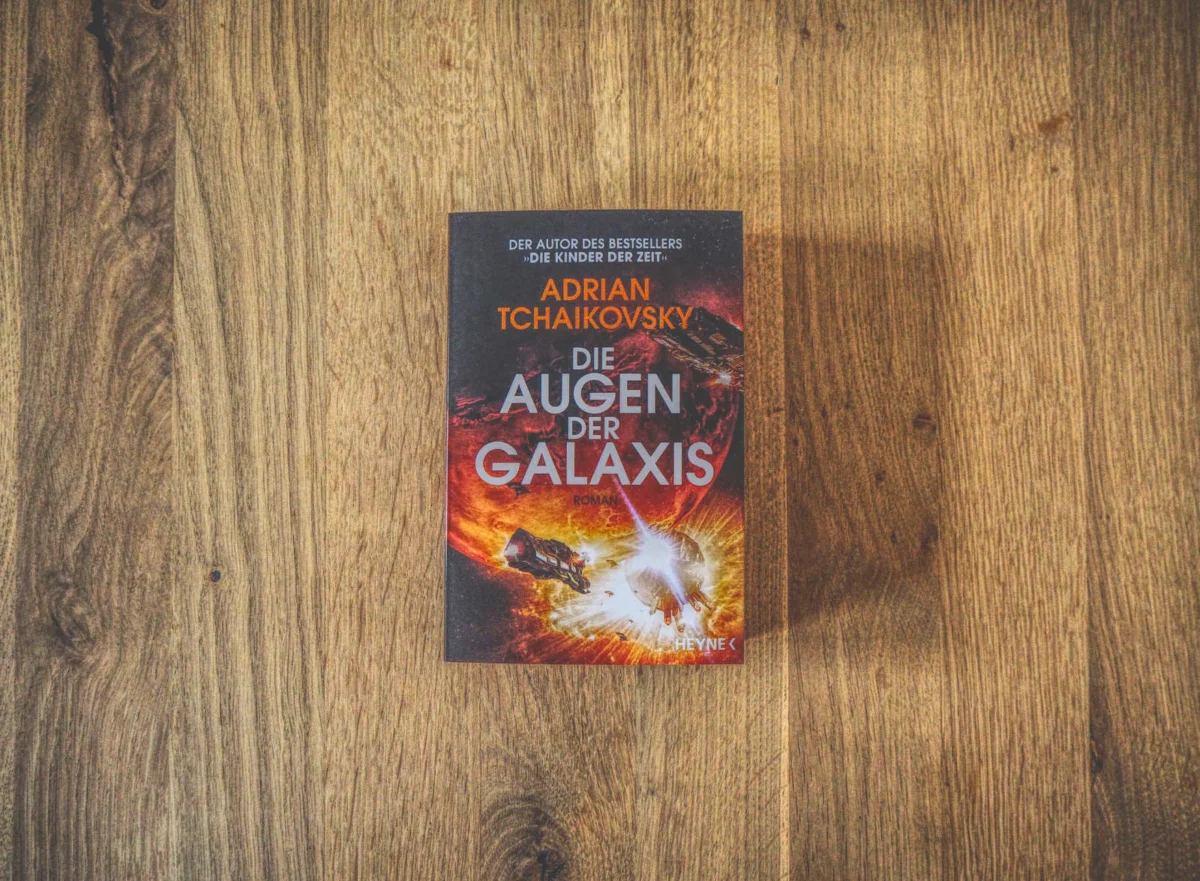 Ein Buch mit dem Titel „Die Augen der Galaxis“ von Adrian Tchaikovsky mit einem Science-Fiction-Cover, platziert auf einer Holzoberfläche.