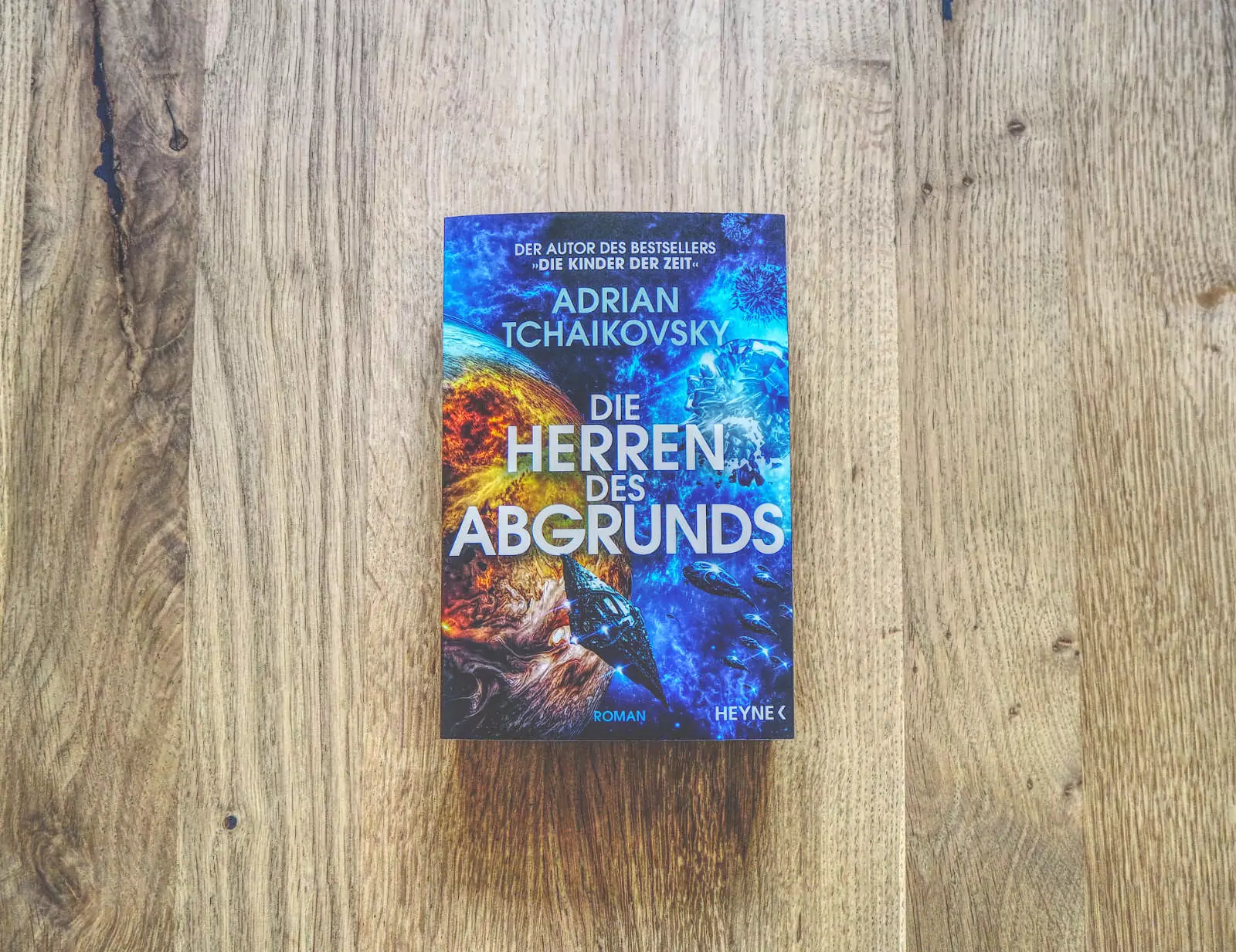 Auf einer Holzfläche liegt eine deutsche Ausgabe des Buches „Die Herren des Abgrunds“ von Adrian Tschaikowsky mit einem farbenfrohen Cover mit kosmischem Thema.