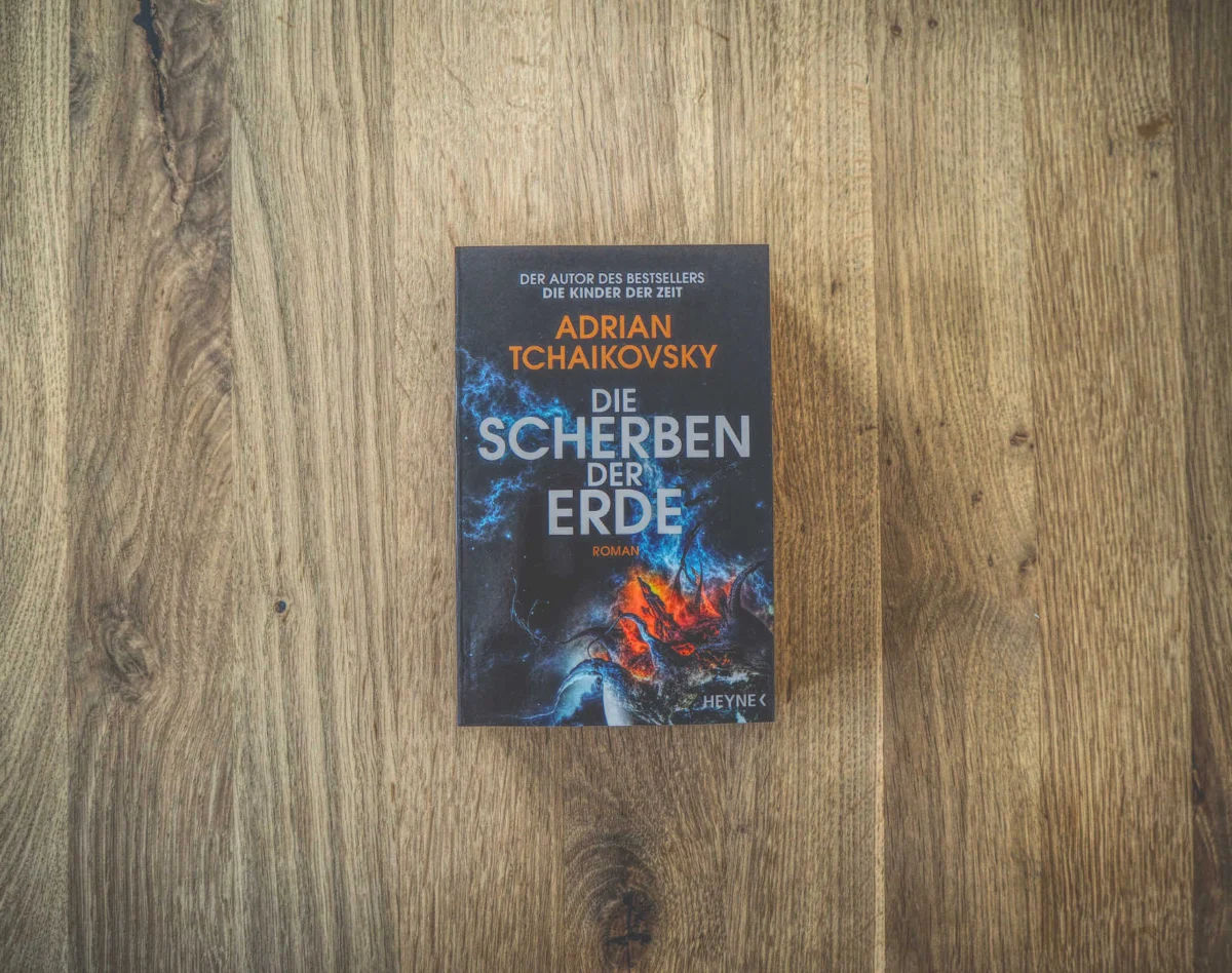Auf einer Holzfläche liegt ein Buch mit dem Titel „Die Scherben der Erde“ von Adrian Tchaikovsky. Der Einband ist mit kosmischen Motiven in Blau und Rot gestaltet.