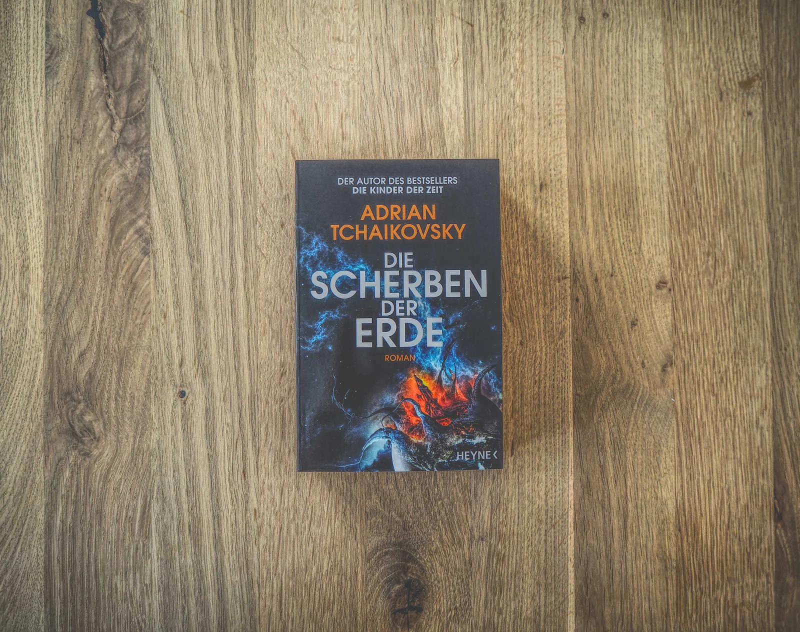 Auf einer Holzfläche liegt ein Buch mit dem Titel „Die Scherben der Erde“ von Adrian Tchaikovsky. Der Einband ist mit kosmischen Motiven in Blau und Rot gestaltet.