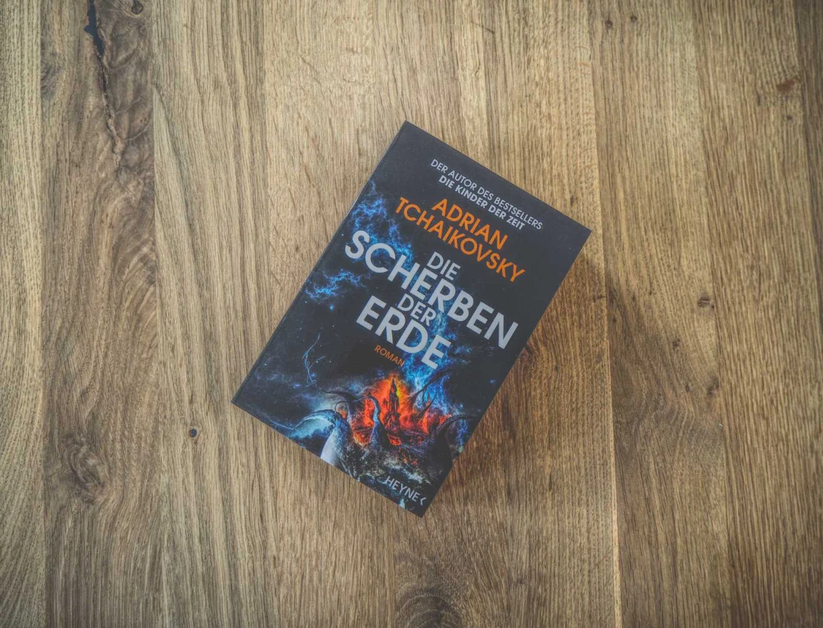 Auf einer Holzunterlage liegt ein Buch mit dem Titel „Die Scherben der Erde“ von Adrian Tchaikovsky.