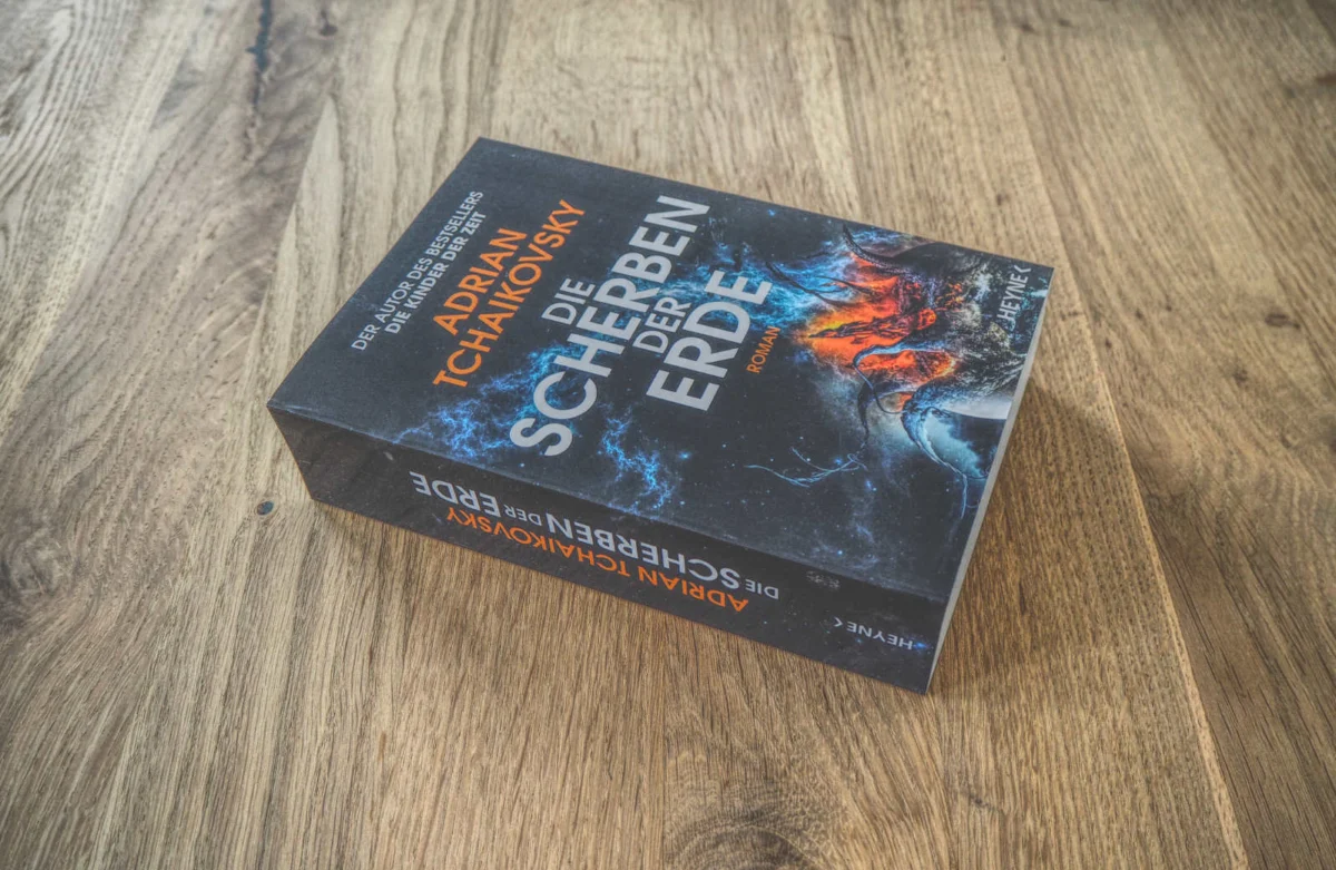 Auf einer Holzfläche liegt ein Buch mit dem Titel „Die Scherben der Erde“ von Adrian Tchaikovsky. Der Einband ist mit feurigen, metallischen Grafiken und einem deutschen Text versehen.