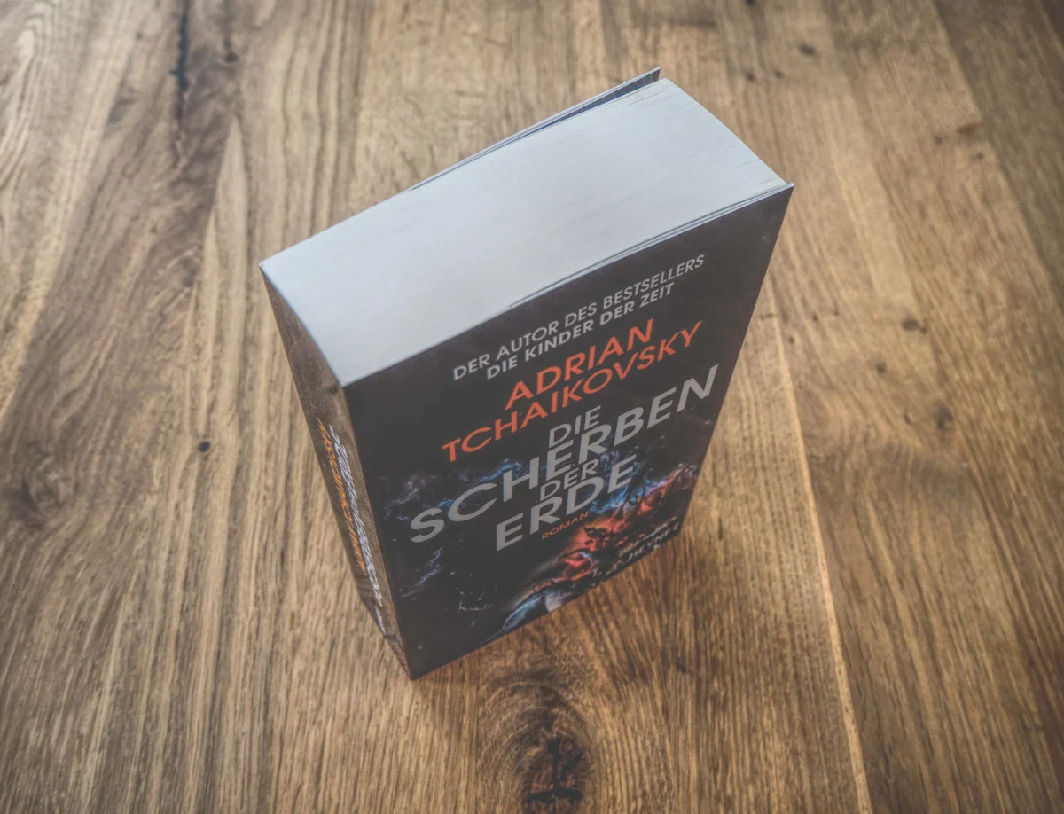 Auf einer Holzfläche steht ein Buch mit dem Titel „Die Scherben der Erde“ von Adrian Tchaikovsky. Der Text ist auf Deutsch.