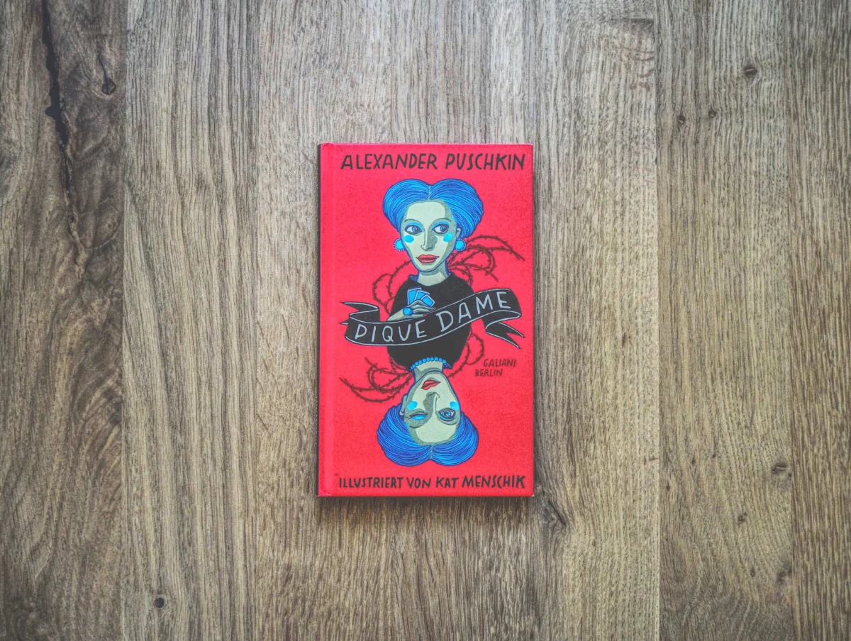 Auf einer Holzfläche liegt ein rot eingebundenes Buch mit dem Titel „Pique Dame“ von Alexander Puschkin, illustriert von Kat Menschik.