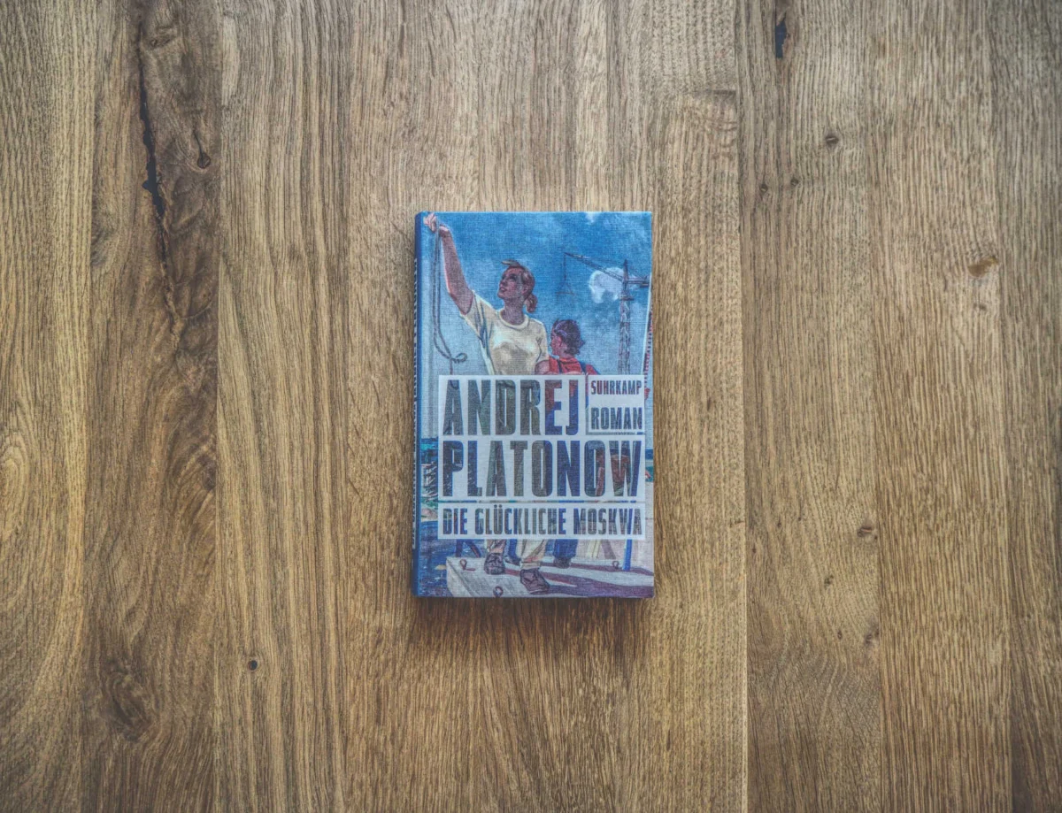 Auf einer Holzfläche liegt ein Buch mit dem Titel "Die glückliche Moskwa" von Andrej Platonow. Auf dem Cover sind Arbeiter und sowjetische Motive abgebildet.