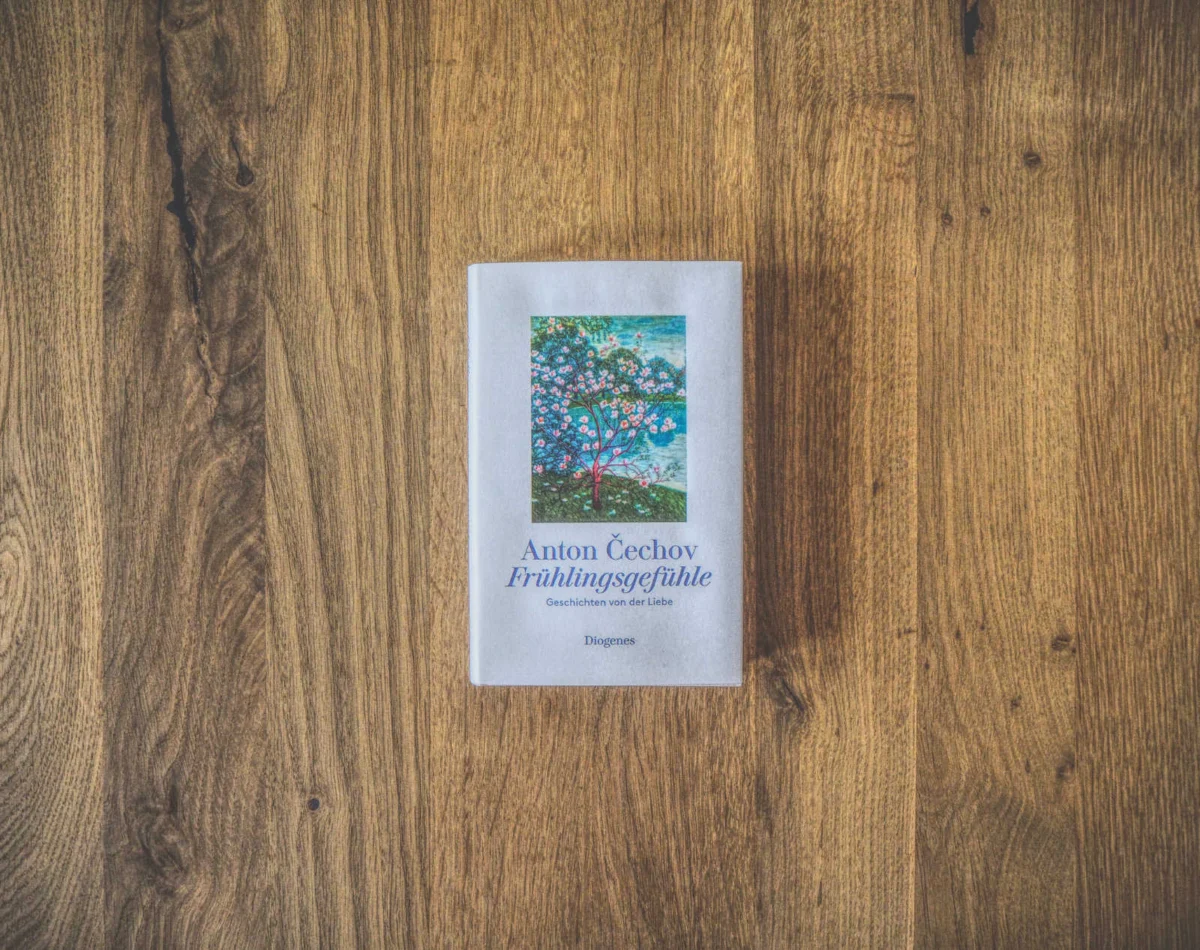 Auf einer Holzfläche liegt ein Buch mit dem Titel „Anton Čechov: Frühlingsgefühle“, auf dessen Cover das Bild eines blühenden Baums zu sehen ist.