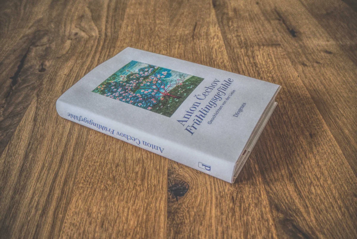 Auf einer Holzfläche liegt ein Buch mit dem Titel „Anton Tschechows Frülingsgefühle“. Auf dem Einband sind blühende Blumen abgebildet.