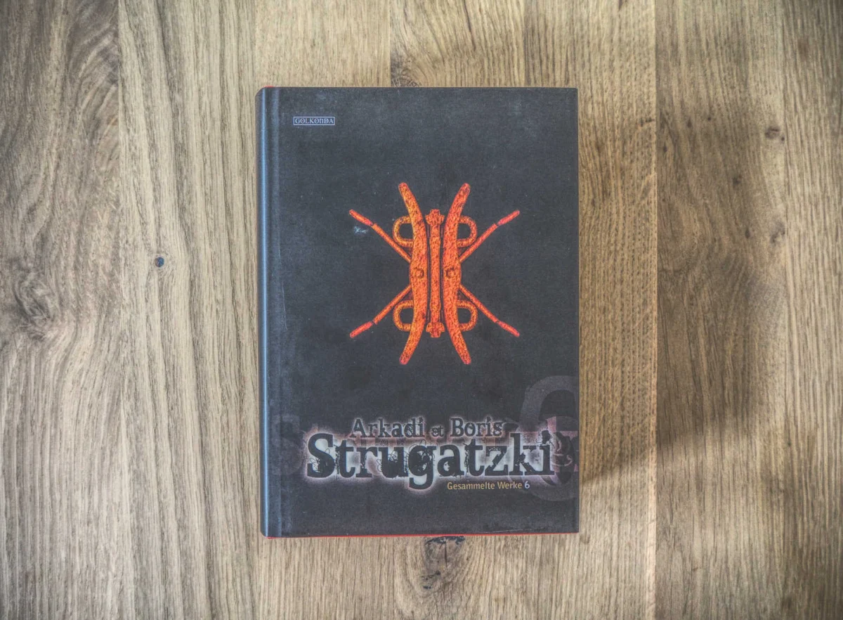 Ein schwarzes Buch mit dem Titel „Strugatzki“ von Arkadi und Boris mit einem detaillierten roten und orangefarbenen Emblem auf dem Cover, platziert auf einer Holzoberfläche.