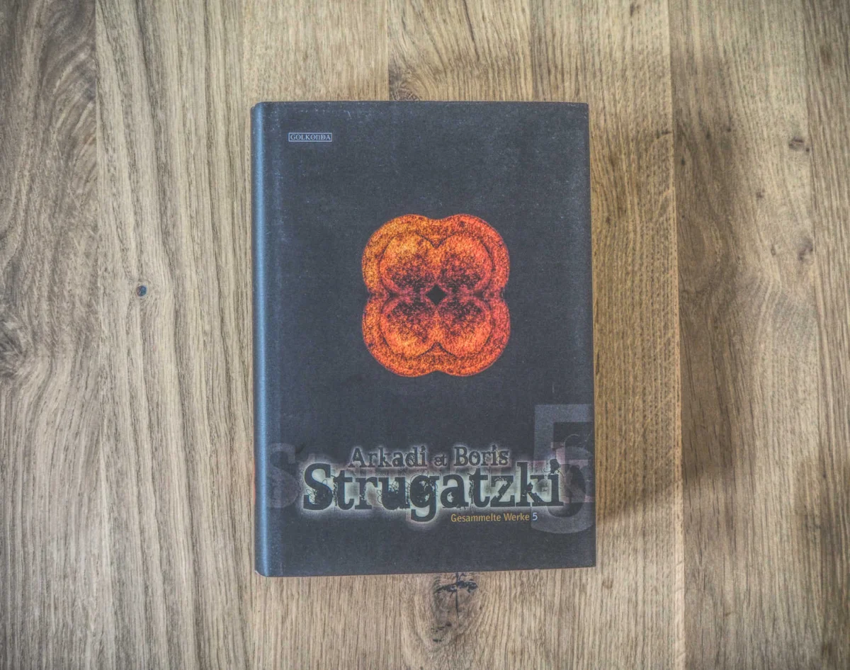 Buch mit dem Titel „Gesammelte Werke 5“ von Arkadi und Boris Strugatzki mit einem dunklen Cover mit einem abstrakten orange-roten Design, platziert auf einer Holzoberfläche.