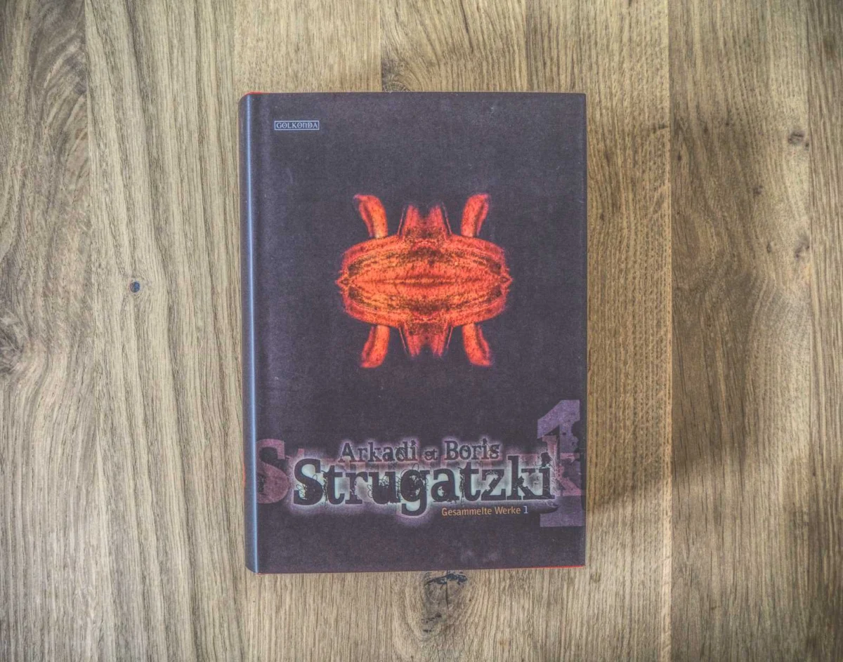 Ein gebundenes Buch mit dem Titel „Arkadi und Boris Strugatzki Gesammelte Werke 1“ mit einem dunklen Einband mit einem abstrakten orange-roten Design, das auf einer Holzoberfläche ruht.