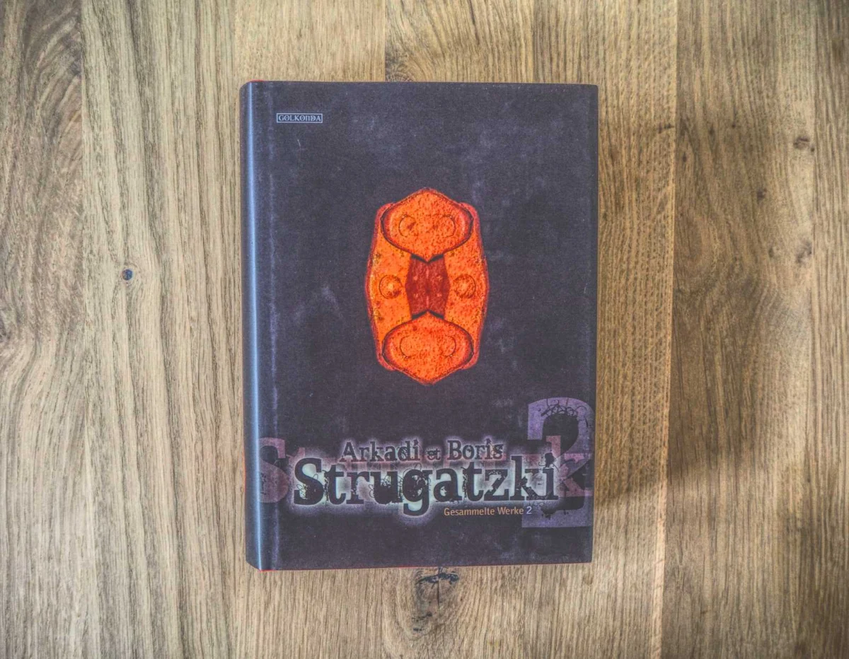 Auf einer Holzfläche liegt ein gebundenes Buch mit dem Titel „Gesammelte Werke 2“ von Arkadi und Boris Strugatzki. Der Einband ist mit abstrakten roten und orangen Mustern verziert.