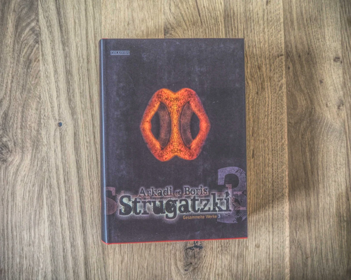 Das Buch mit dem Titel „Arkadi und Boris Strugatzki Gesammelte Werke 3“ liegt auf einer Holzoberfläche. Der Einband zeigt rot-orange abstrakte Formen.