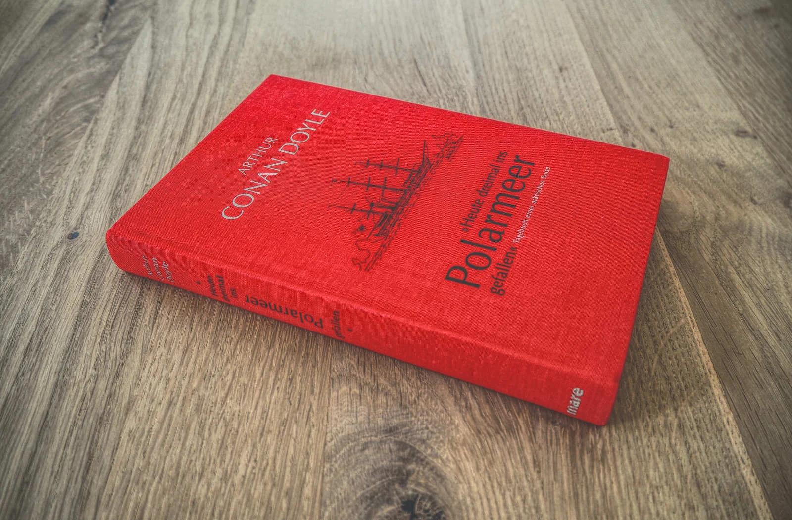 Ein rotes gebundenes Buch mit dem Titel „Heute dreimal ins Polarmeer gefallen“ von Arthur Conan Doyle liegt auf einer Holzoberfläche.