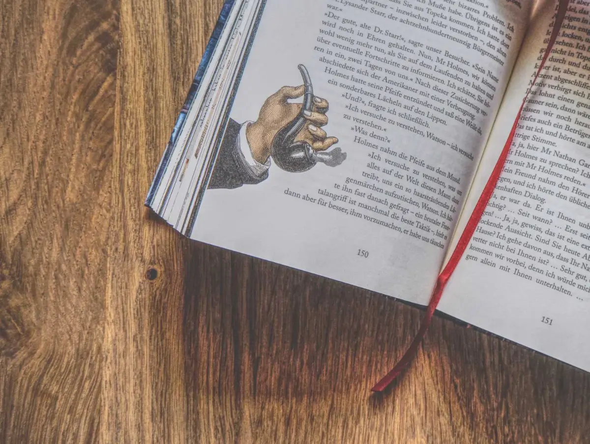 Ein aufgeschlagenes Buch mit der Illustration einer Hand, die eine Pfeife hält, ist auf einer hölzernen Oberfläche abgebildet, wobei ein Lesezeichen mit rotem Band sichtbar ist.
