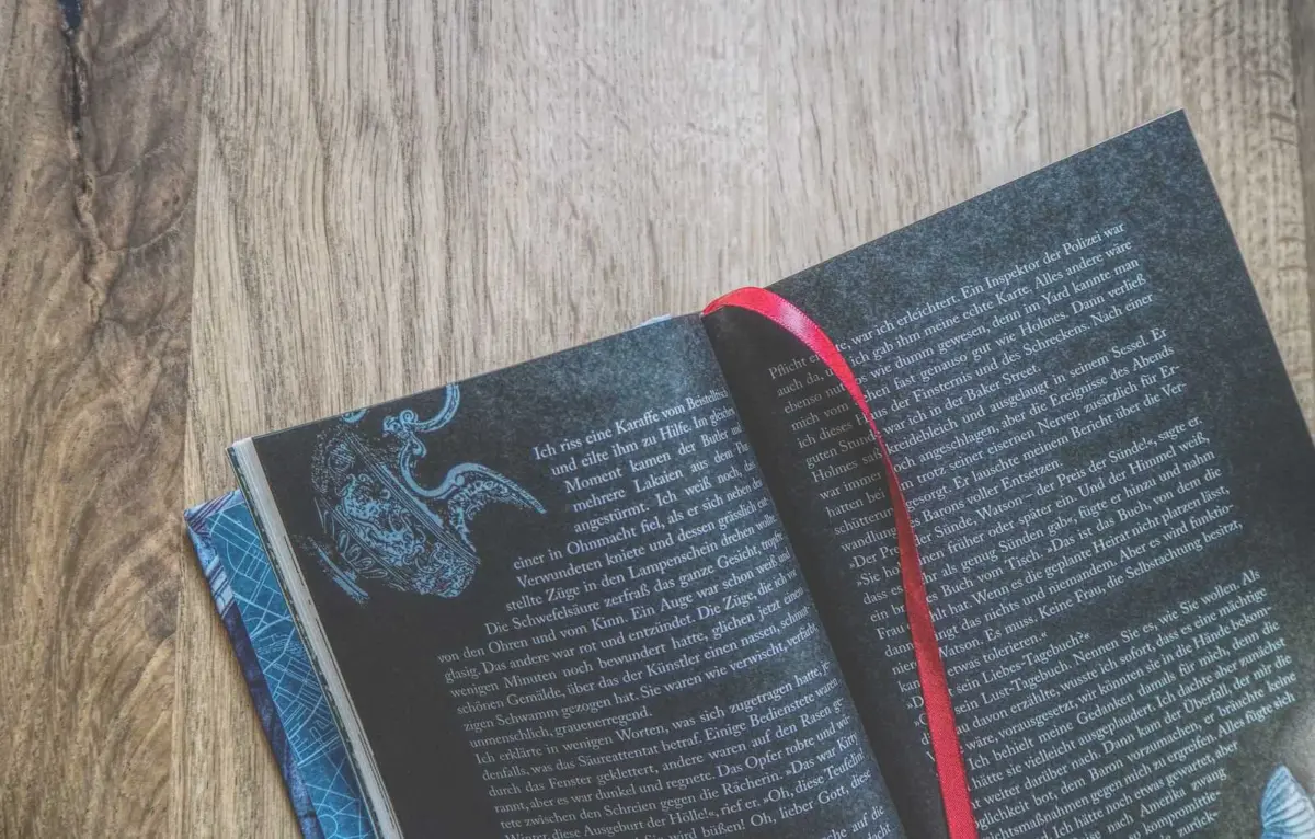Ein aufgeschlagenes Buch mit weißem deutschem Text auf dunklen Seiten, einem Lesezeichen mit rotem Band und einem Teil des blauen Einbands, das auf einer Holzfläche liegt.