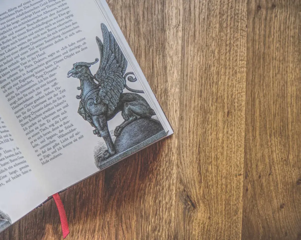 Ein aufgeschlagenes Buch mit einer Schwarz-Weiß-Illustration eines geflügelten Greifs auf einer hölzernen Oberfläche; der Text ist auf Deutsch.