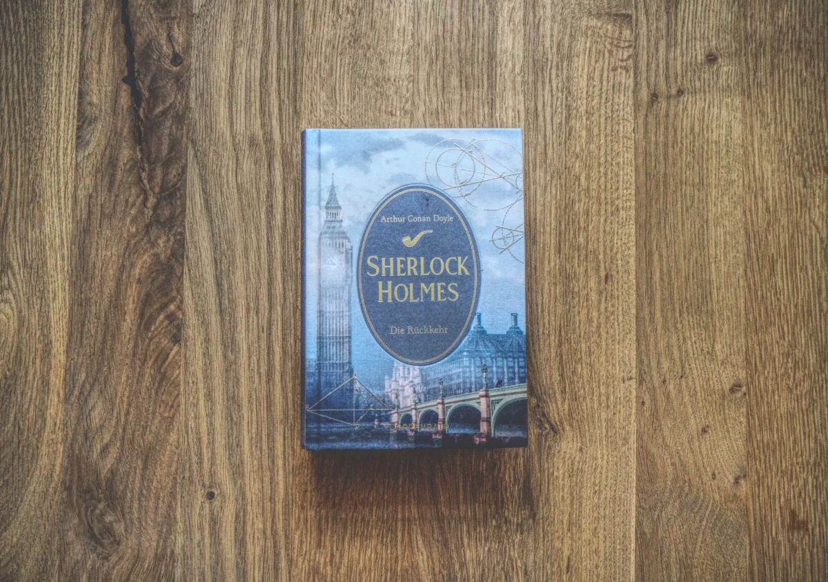 Auf einer Holzfläche liegt ein gebundenes Buch mit dem Titel „Sherlock Holmes: Die Rückkehr“ von Arthur Conan Doyle. Auf dem Cover sind Abbildungen von Londoner Sehenswürdigkeiten zu sehen.