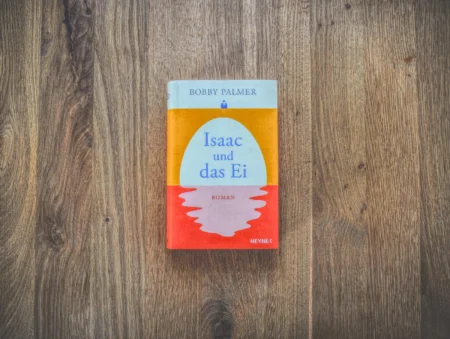 Auf einer Holzfläche liegt ein deutsches Buch mit dem Titel „Isaac und das Ei“ von Bobby Palmer.