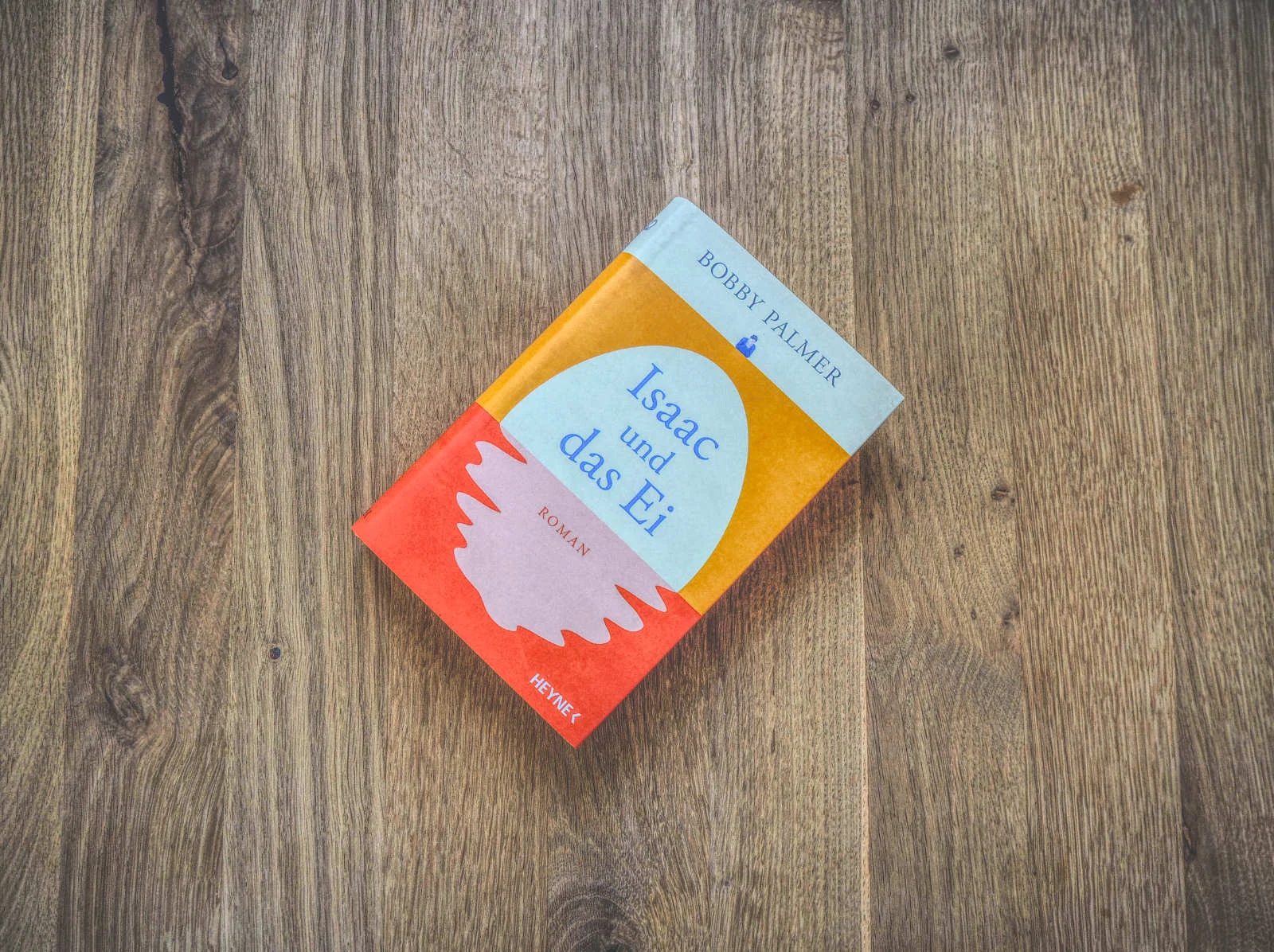 Auf einer Holzoberfläche liegt ein Buch mit dem Titel „Isaac und das Ei“ von Bobby Palmer. Der Einband ist mit einem abstrakten Design in Blau-, Orange- und Rottönen gestaltet.