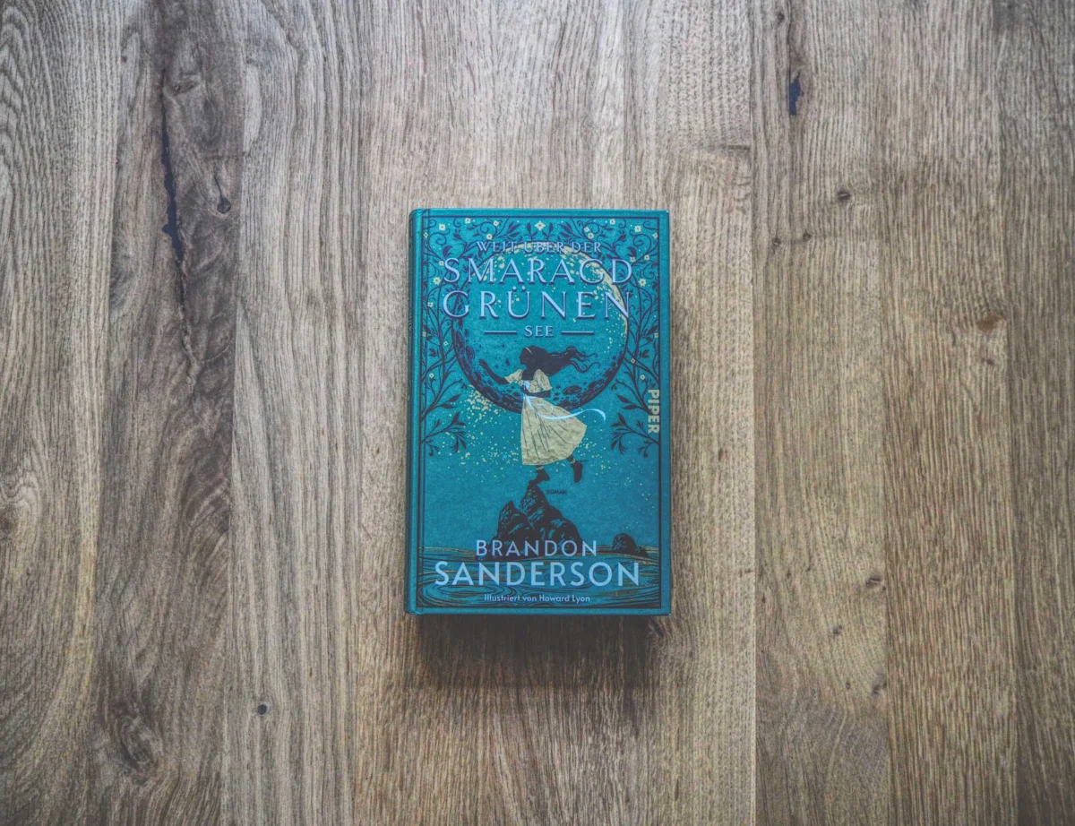 Ein gebundenes Buch mit dem Titel „Weit über der smaragdgrünen See“ von Brandon Sanderson, auf einer Holzoberfläche platziert. Der Buchumschlag zeigt aufwendige grüne und blaue Illustrationen.