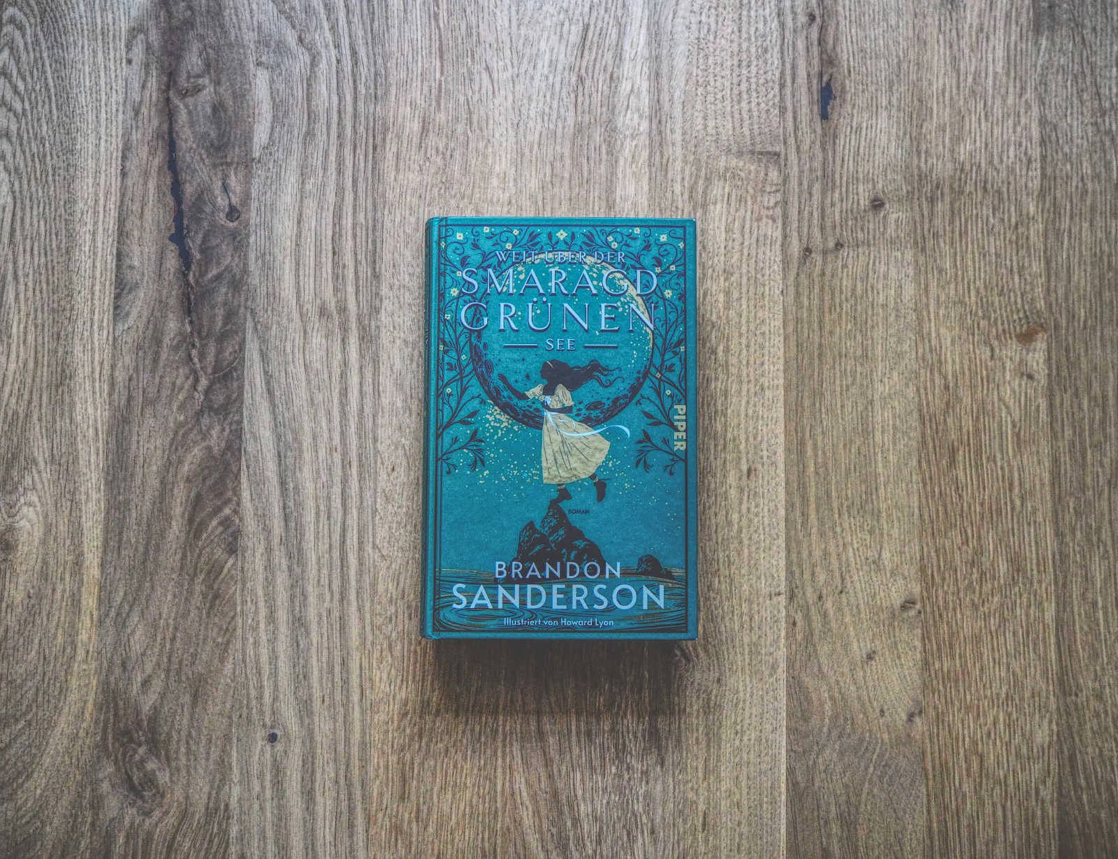 Ein gebundenes Buch mit dem Titel „Weit über der smaragdgrünen See“ von Brandon Sanderson, auf einer Holzoberfläche platziert. Der Buchumschlag zeigt aufwendige grüne und blaue Illustrationen.