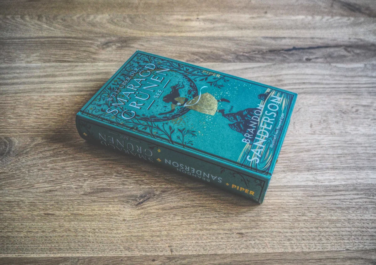 Ein gebundenes Buch mit dem Titel „Weit über der smaragdgrünen See“ von Brandon Sanderson liegt mit der Vorderseite nach oben auf einer Holzoberfläche. Der Buchumschlag ist überwiegend blaugrün mit goldenen und schwarzen Akzenten.