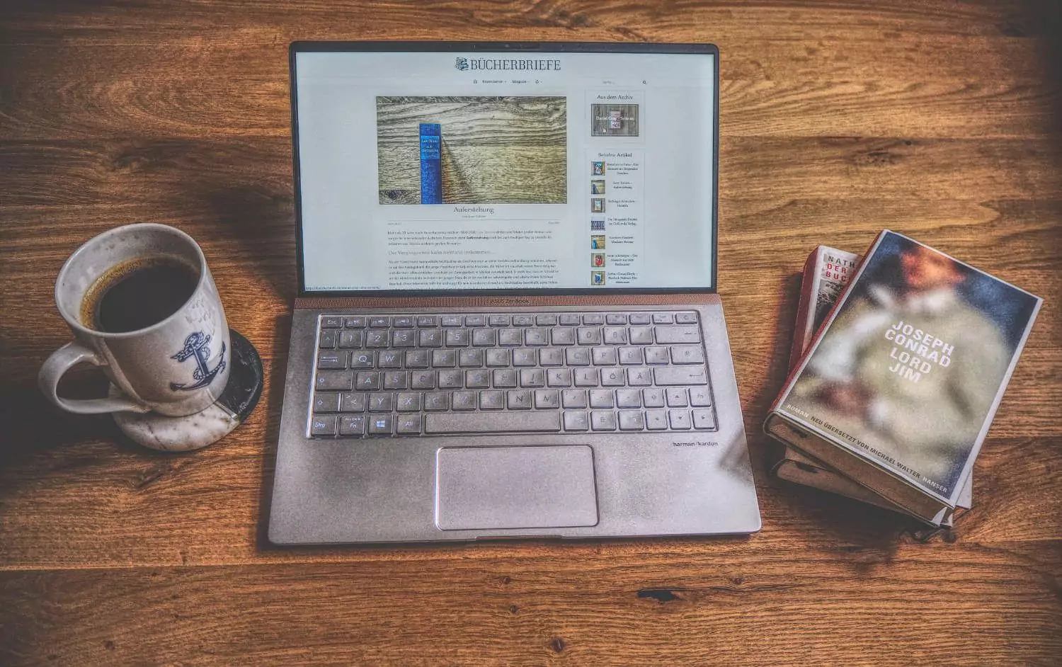 Ein Laptop auf einem Holztisch neben einem Buch und einer Tasse Kaffee.