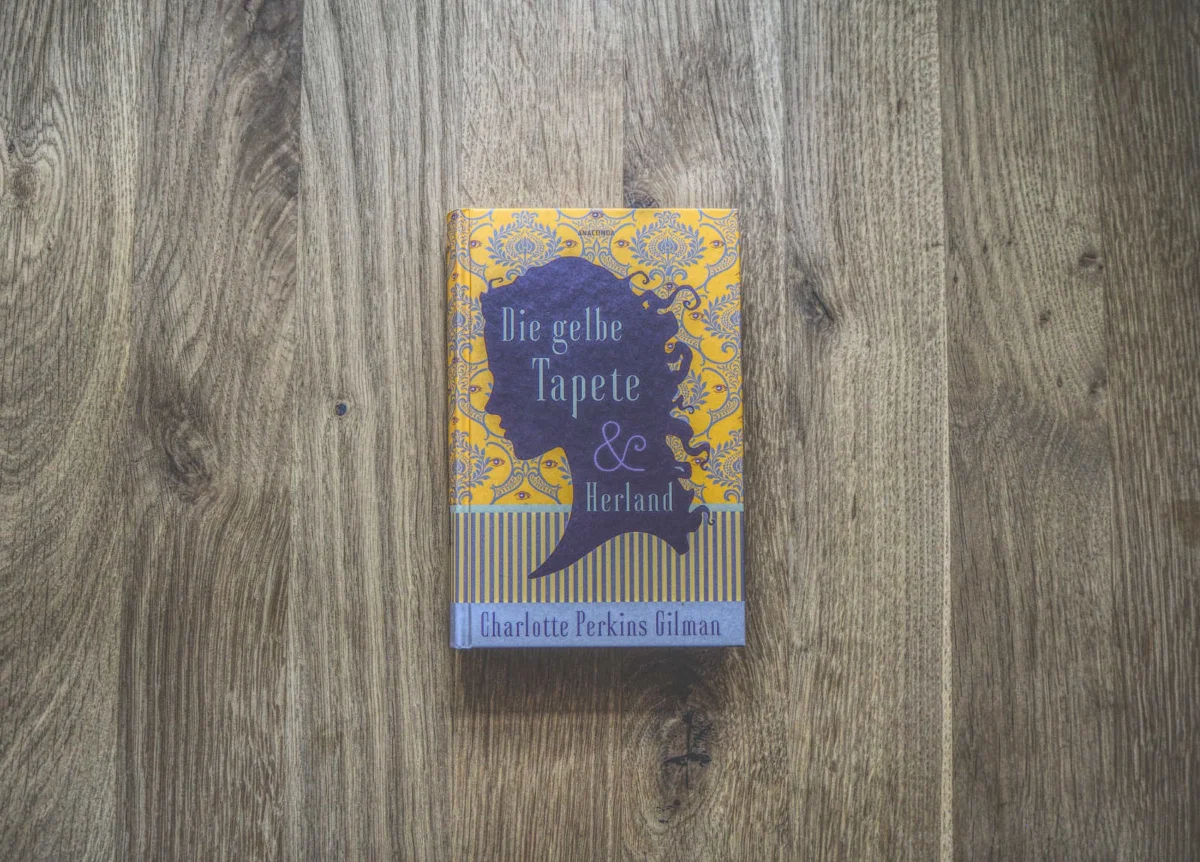 Auf einer Holzoberfläche liegt ein Buch mit dem Titel „Die gelbe Tapete & Herland“ von Charlotte Perkins Gilman, mit einem gelb-violetten Einband, der ein Blumenmuster und eine weibliche Silhouette zeigt.