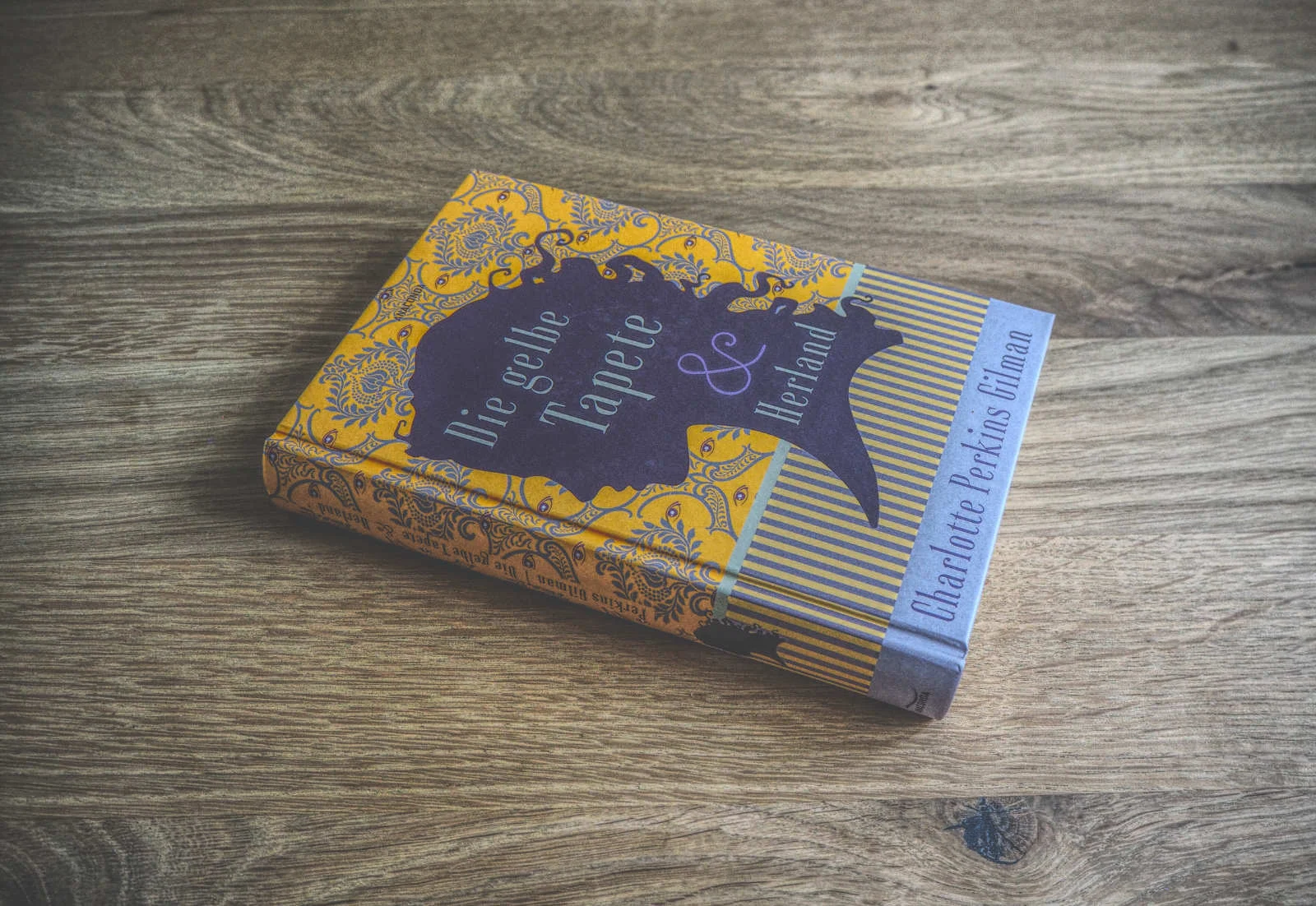 Ein gebundenes Buch mit dem Titel „Die gelbe Tapete“ von Charlotte Perkins Gilman auf einer Holzoberfläche. Der Einband ist mit einem kunstvollen gelb-violetten Design versehen.