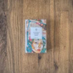 Ein Buch mit dem Titel „Ich und Jimmy“ von Clarice Lispector mit einem farbenfrohen, abstrakten Cover mit einem Gesicht und verschiedenen Mustern, platziert auf einer Holzoberfläche.