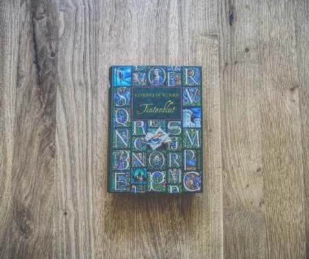 Auf einer Holzfläche liegt ein Buch mit dem Titel „Tintenblut“ von Cornelia Funke. Das Buch hat einen verzierten Einband mit bunten Illustrationen und verzierten Buchstaben.