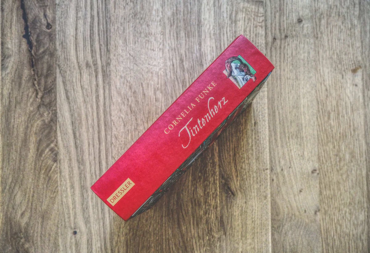 Auf einer Holzplatte liegt ein rotes Buch von Cornelia Funke mit dem Titel „Tintenherz“. Auf dem Buchrücken sind der Name der Autorin, der Titel und der Verlag Dressler zu sehen.