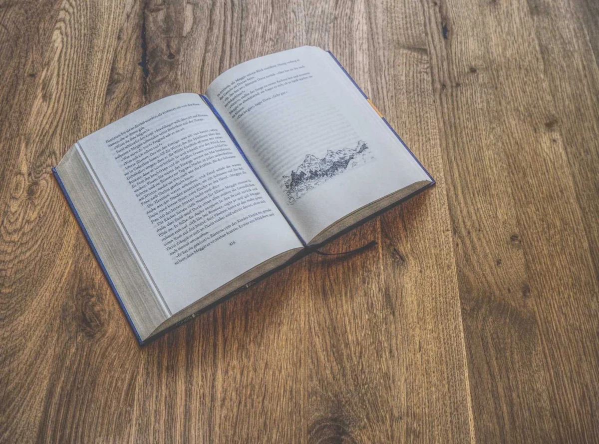 Auf einem Holztisch liegt ein offenes Buch mit Text und einer Bergillustration.