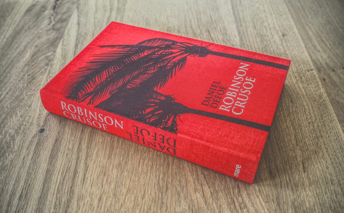 Auf einer Holzfläche liegt ein rotes Hardcover-Buch mit dem Titel „Robinson Crusoe“ von Daniel Defoe. Auf dem Cover sind die schwarze Silhouette eines Seemanns und Palmen zu sehen.