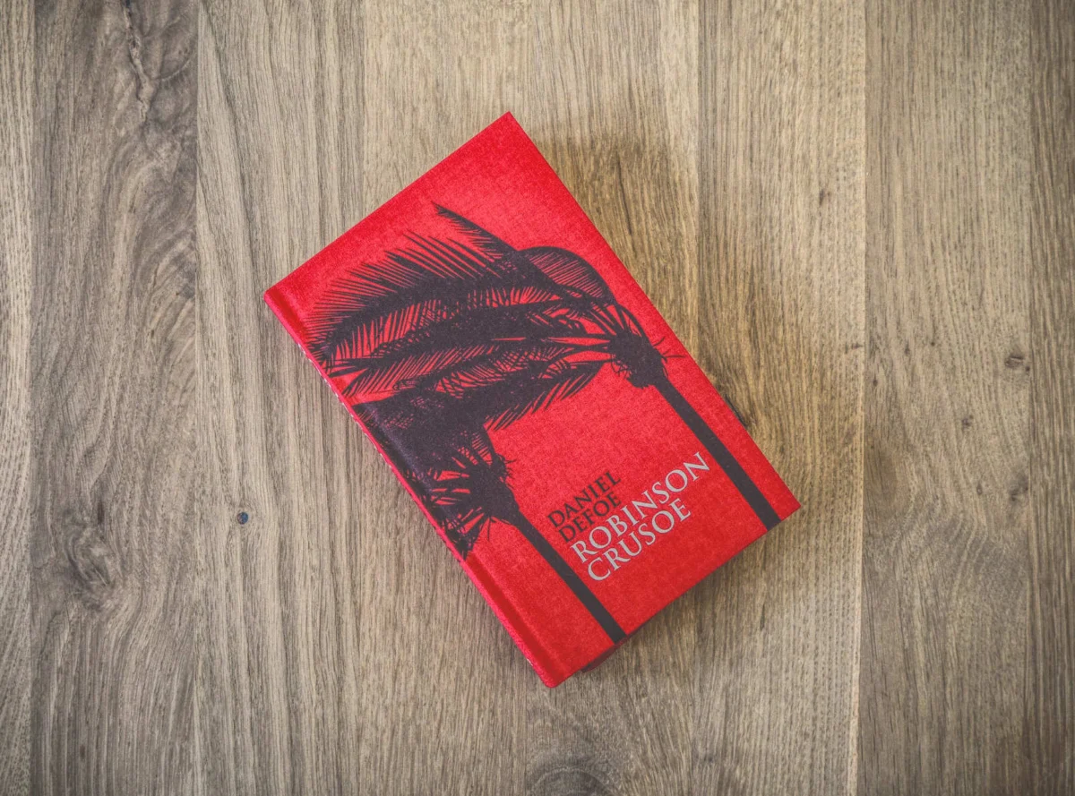 Ein rotes gebundenes Buch mit dem Titel „Robinson Crusoe“ von Daniel Defoe. Auf dem Cover ist die schwarze Silhouette einer Palme zu sehen, platziert auf einer Holzoberfläche.