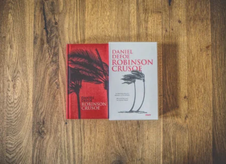 Zwei Ausgaben des Buches „Robinson Crusoe“ von Daniel Defoe liegen nebeneinander auf einer Holzfläche. Ein Buch hat einen roten Einband mit schwarzen Palmen, das andere einen beigen Einband mit schwarzen Palmen.