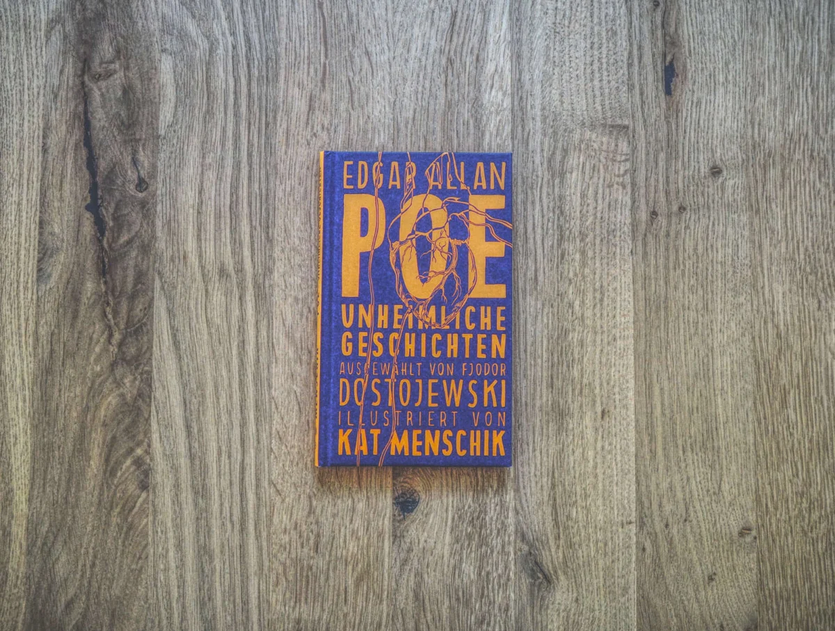 Auf einer Holzunterlage liegt ein Buch mit deutschem Text auf dem Cover, betitelt „Edgar Allan Poe: Unheimliche Geschichten“, illustriert von Kat Menschik.