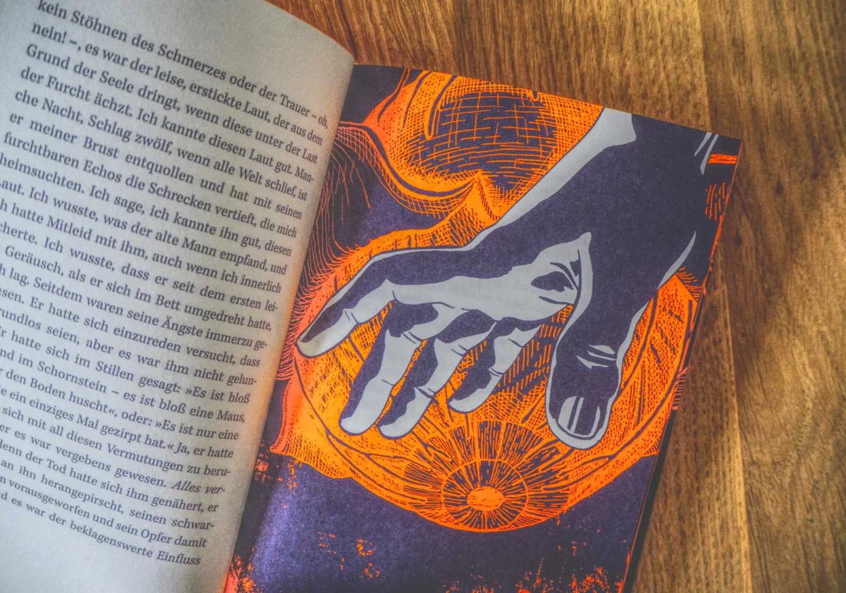 Aufgeschlagenes Buch auf Holzuntergrund mit orange-blauer Hand-Illustration auf der einen Seite und deutschem Text auf der gegenüberliegenden Seite.