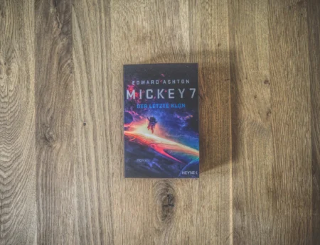 Buch mit dem Titel „Mickey7: Der Letzte Klon“ von Edward Ashton auf einer Holzoberfläche. Das Cover zeigt einen Astronauten, der im farbenfrohen Weltraum schwebt.
