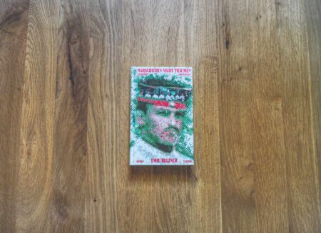 Ein Buch, auf dessen Einband das Gesicht eines Mannes zu sehen ist, liegt auf einer hellbraunen Holzfläche. Der Titel des Buches ist in einer fremden Sprache verfasst und in roter und grüner Schrift geschrieben.