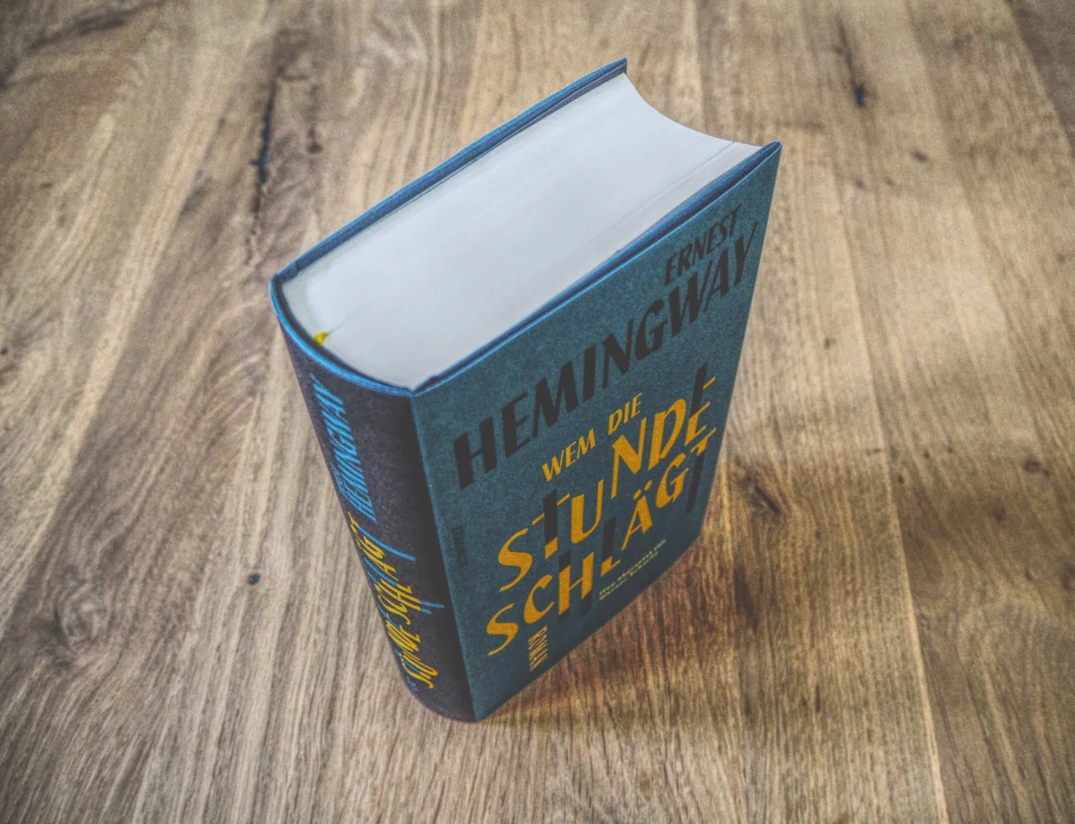 Ein dickes gebundenes Buch mit dem Titel „Wem die Stunde schlägt“ von Ernest Hemingway, aufrecht auf einer Holzoberfläche platziert.