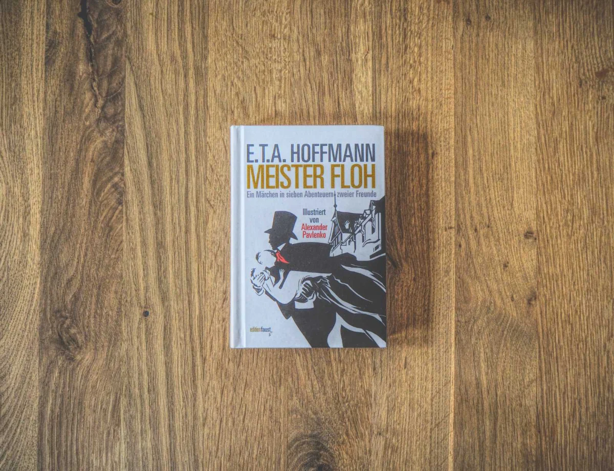 Auf einer Holzfläche liegt ein gebundenes Buch mit dem Titel „E.T.A. Hoffmann -Meister Floh“ und illustriertem Cover.
