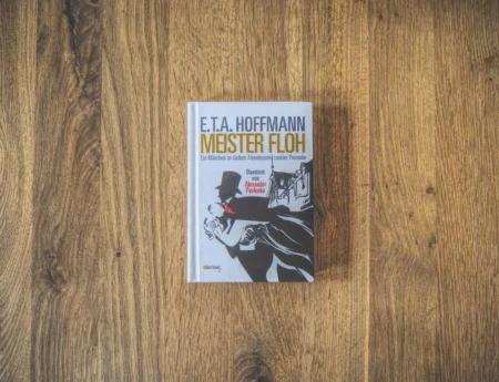Auf einer Holzfläche liegt ein gebundenes Buch mit dem Titel „E.T.A. Hoffmann -Meister Floh“ und illustriertem Cover.