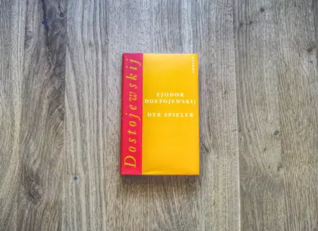 Auf einer Holzfläche liegt ein Buch mit gelbem Einband und dem Titel „Der Spieler“ von Fjodor Dostojewskij.