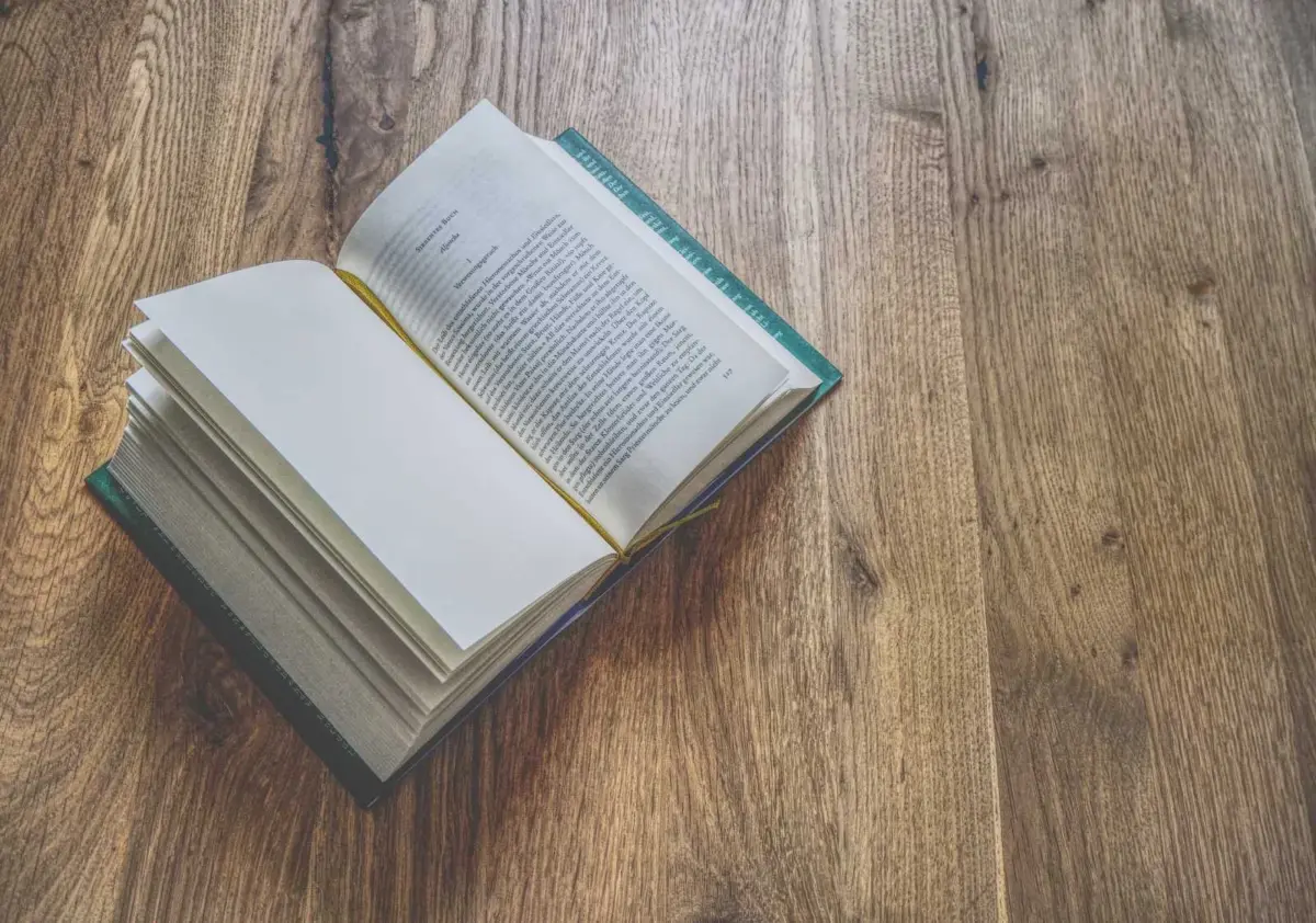Ein aufgeschlagenes Buch mit sichtbarem Text liegt auf einer hölzernen Unterlage, auf der mehrere Seiten nach links und rechts umgeschlagen sind.
